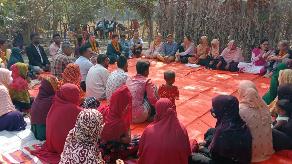 Large group of people sit on red rugs around a square in conversation