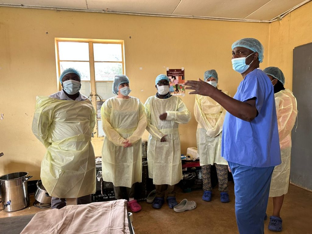 A photo of 6 people stood in an operating room, wearing hospital overalls and masks. 