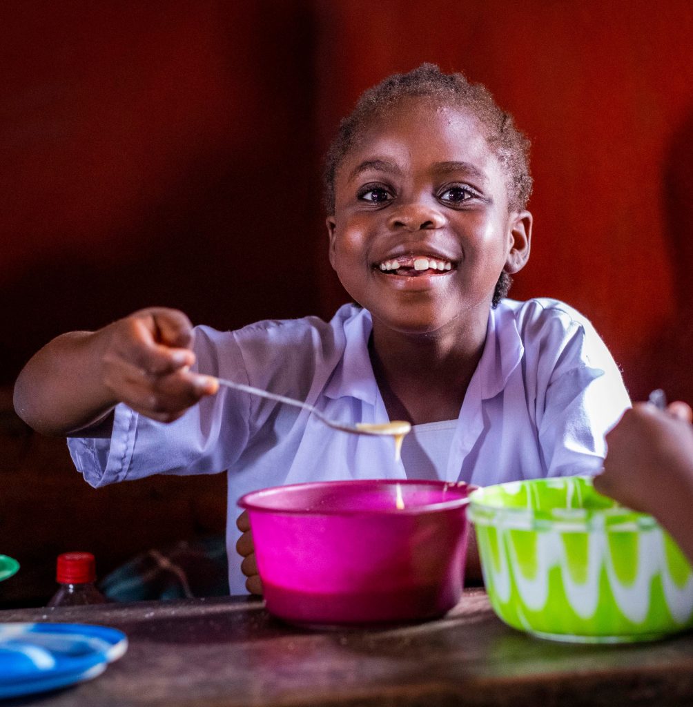 A girl sat at a table eating some food from a bowl, holding up a spoon.