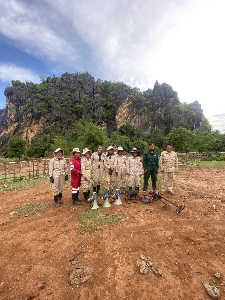 A group of people stood on a piece of land wearing overalls and hats with some clearing eqipment.