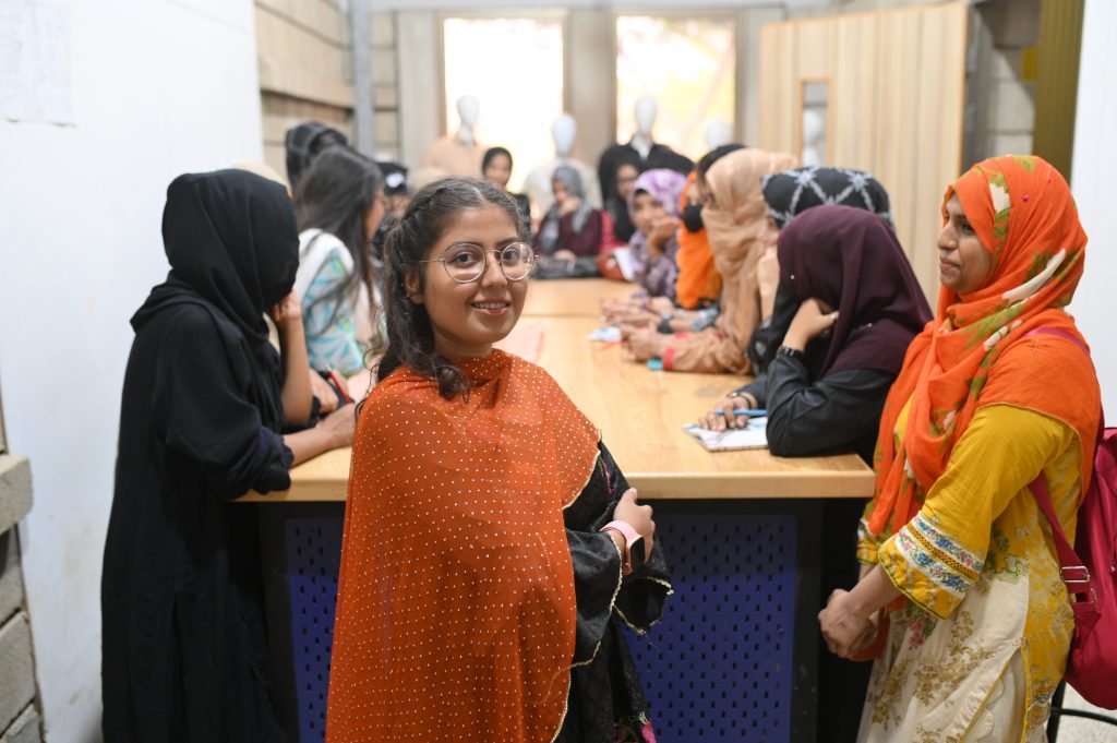 A group of women sat around a table wearing colour clothing. A woman stood at the front with her arms crossed, smiling. 