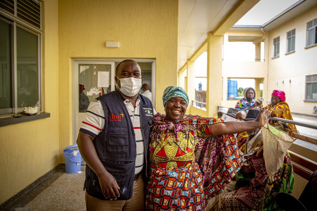 A woman in brightly coloured clothes has one arm around the doctor, the other arm raised in celebration, a smile beaming on her face. 