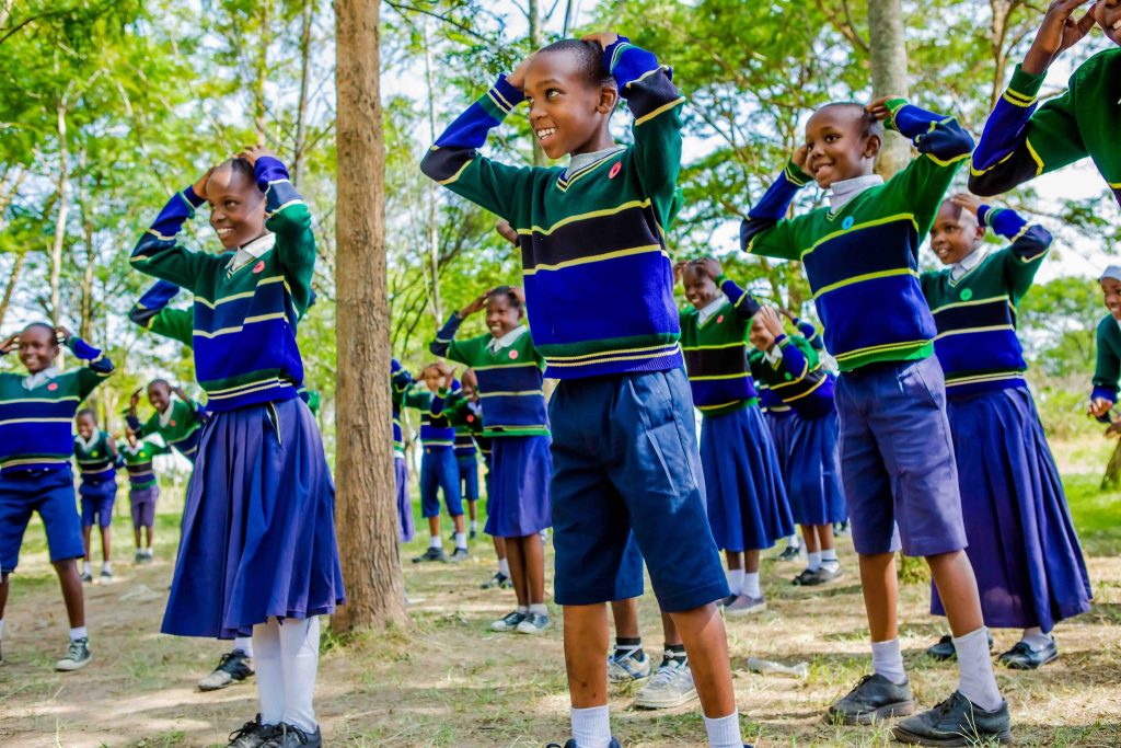 Children stood with their hands on their head playing a game, wearing green and blue striped school uniforms, amongst a backdrop of trees