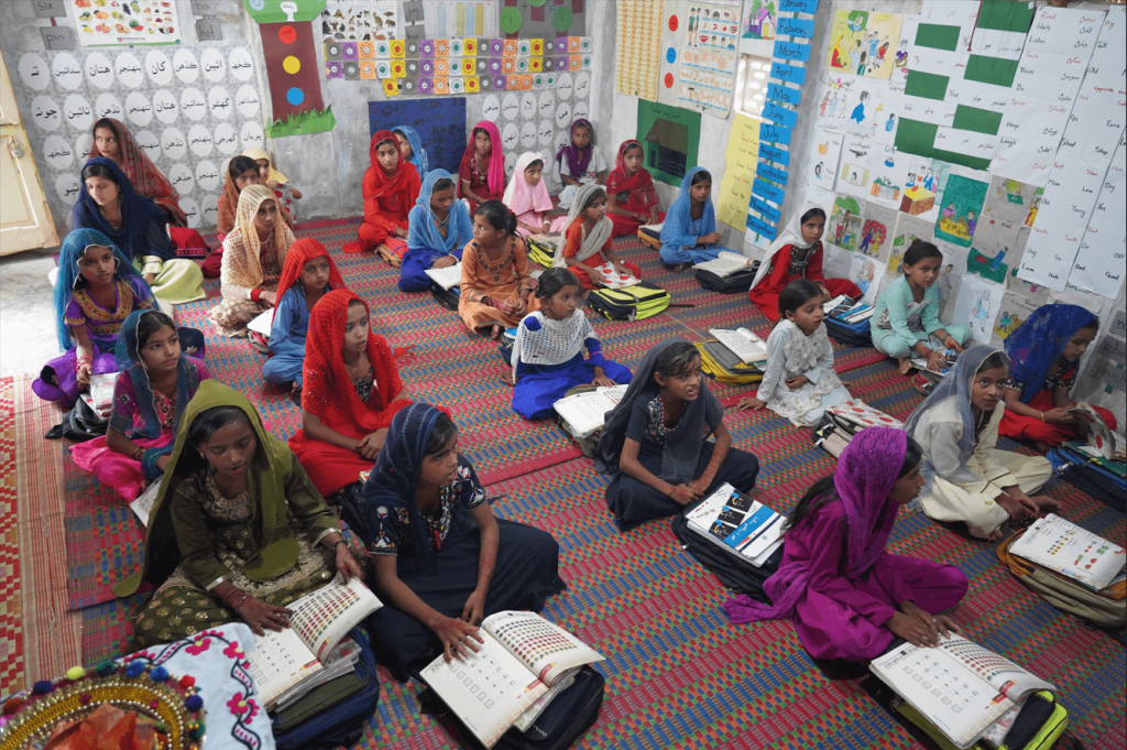 Girls in colourful clothing sat on a rug on the floor with books open, and learning materials all over the walls.